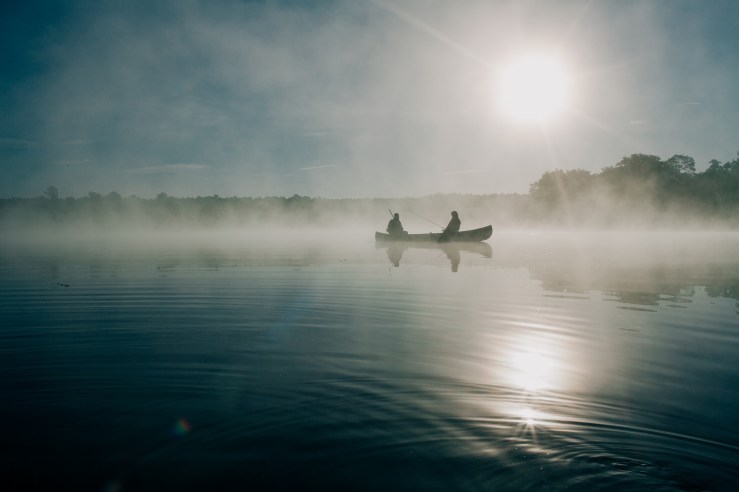 Two people are in a canoe in the early morning. Mist is over the water and trees can be seen on the horizon. Near the top right of the boat is the rising sun. Its reflection is visible on the water.