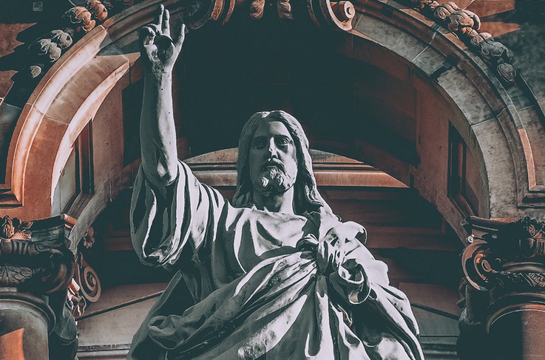 A sculpture of Jesus. His right hand is raised and He is looking ahead. He looks like He is in control. He is wearing a tunic. An arch with columns can be seen behind Him.