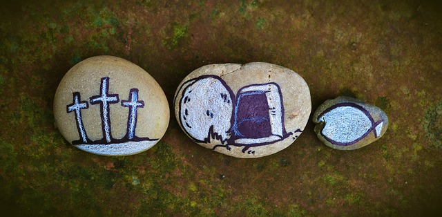 Three rocks on the ground that are painted on. The first shows three crosses. the second shows an empty tomb. The third is smaller than the other two, and has a "Jesus fish" drawn on it.