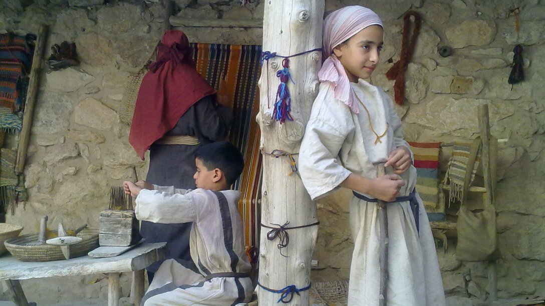 Two young boys in Israel weaving in front of their home. There is a pole in the middle of the image with one of the boys leaning up against it. Another boy on the left is sitting down and working.