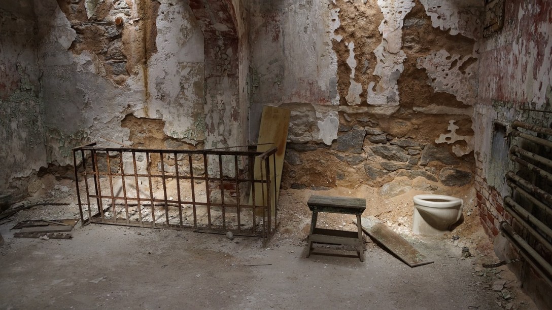 A ruined prison cell. Almost half of the walls don't have paint on them, and the floor is just dirt. There is a bed frame, but it is leaning up against a wall. There is a toilet in the corner, with no way to flush it.