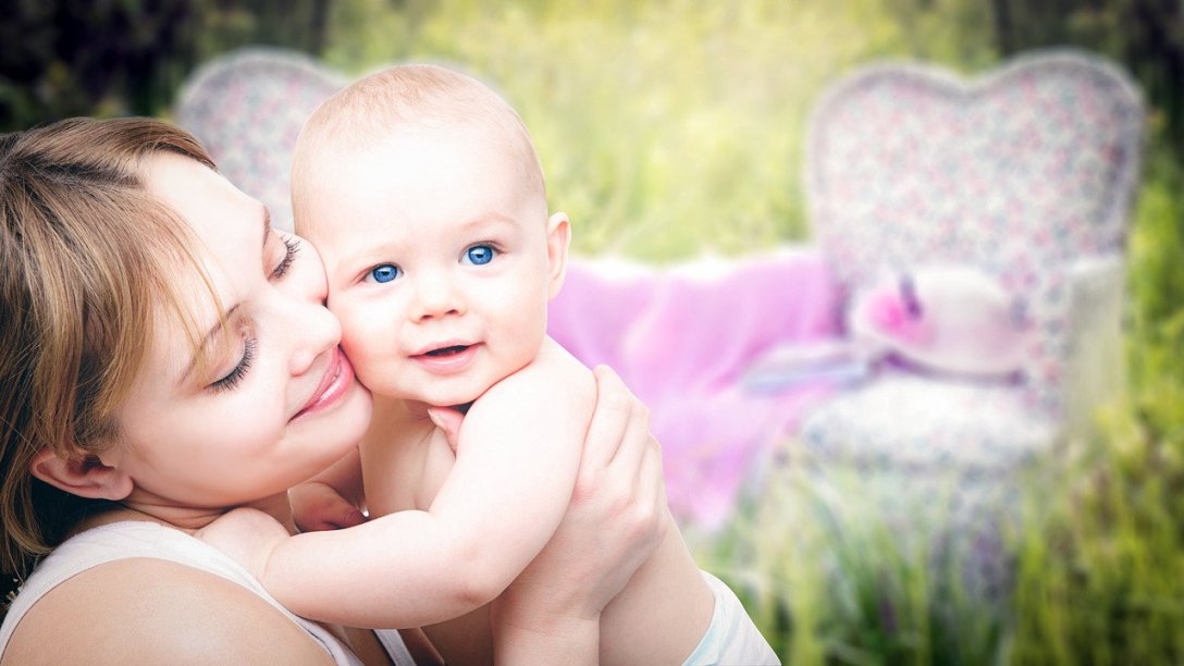 A mother with her baby. She has a smile on her face, and is pressing her cheek up against her baby's. The has bright, blue eyes, and is looking into the distance. A couple of large chairs can be seen in the background.
