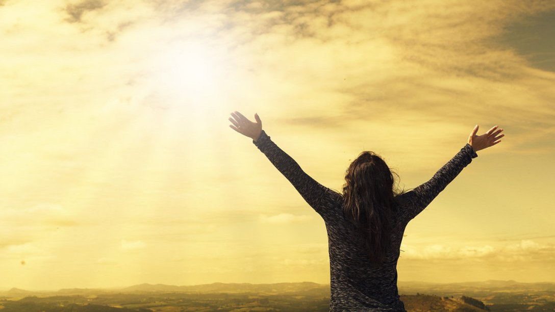 A woman looking up at the sun in the sky. Her arms are outstretched in an expression of praise.