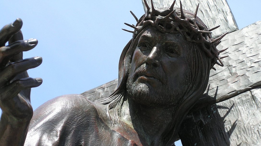 A close-up of a sculpture showing Jesus carrying His cross. In the photo, His cross is over His left shoulder and He is looking toward the right. His right hand is near eye level and on the left side of the image.