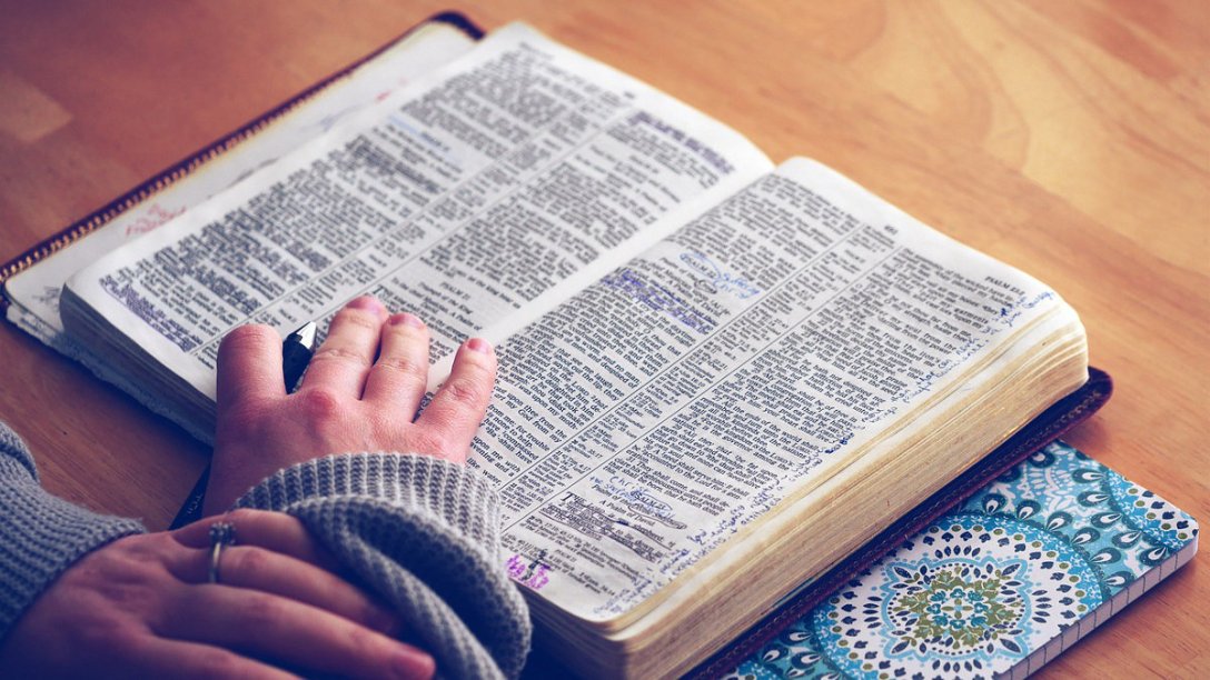 A Bible is open to the book of Psalms. A person has his or her hand on it to keep it open, with a pen tucked between two fingers. He or she is reading, but also ready to make notes in the margins as needed.